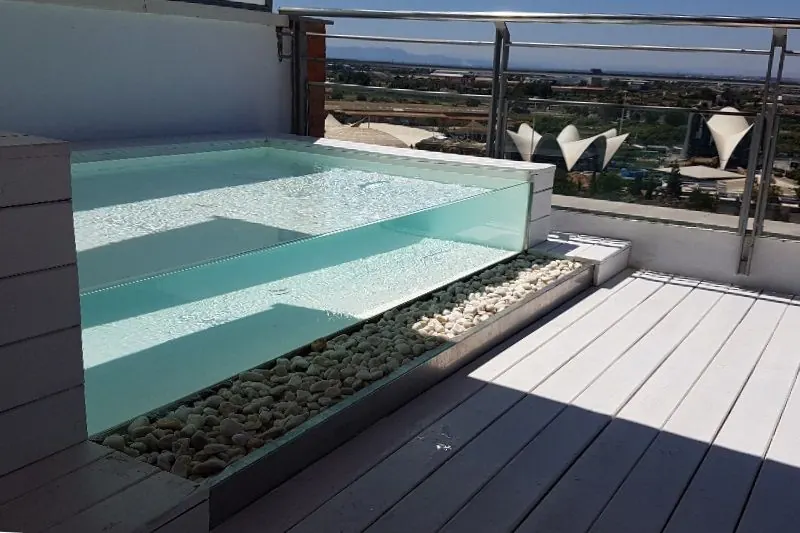 La piscina de vidrio se apoya sobre un lecho de canto rodado, de modo que el agua queda a la vista desde la tarima. Al fondo, las cubiertas blancas del Ágora.