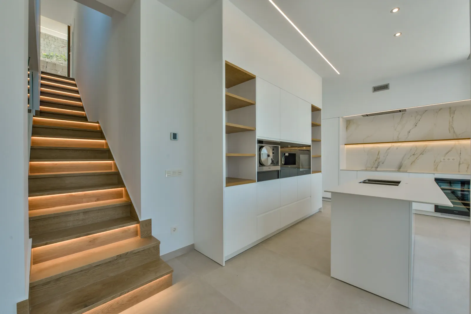 La escalera de roble, con luz continua bajo cada peldaño, separa el acceso de la cocina, resuelta en un frente compacto con hornos empotrados y baldas de madera.