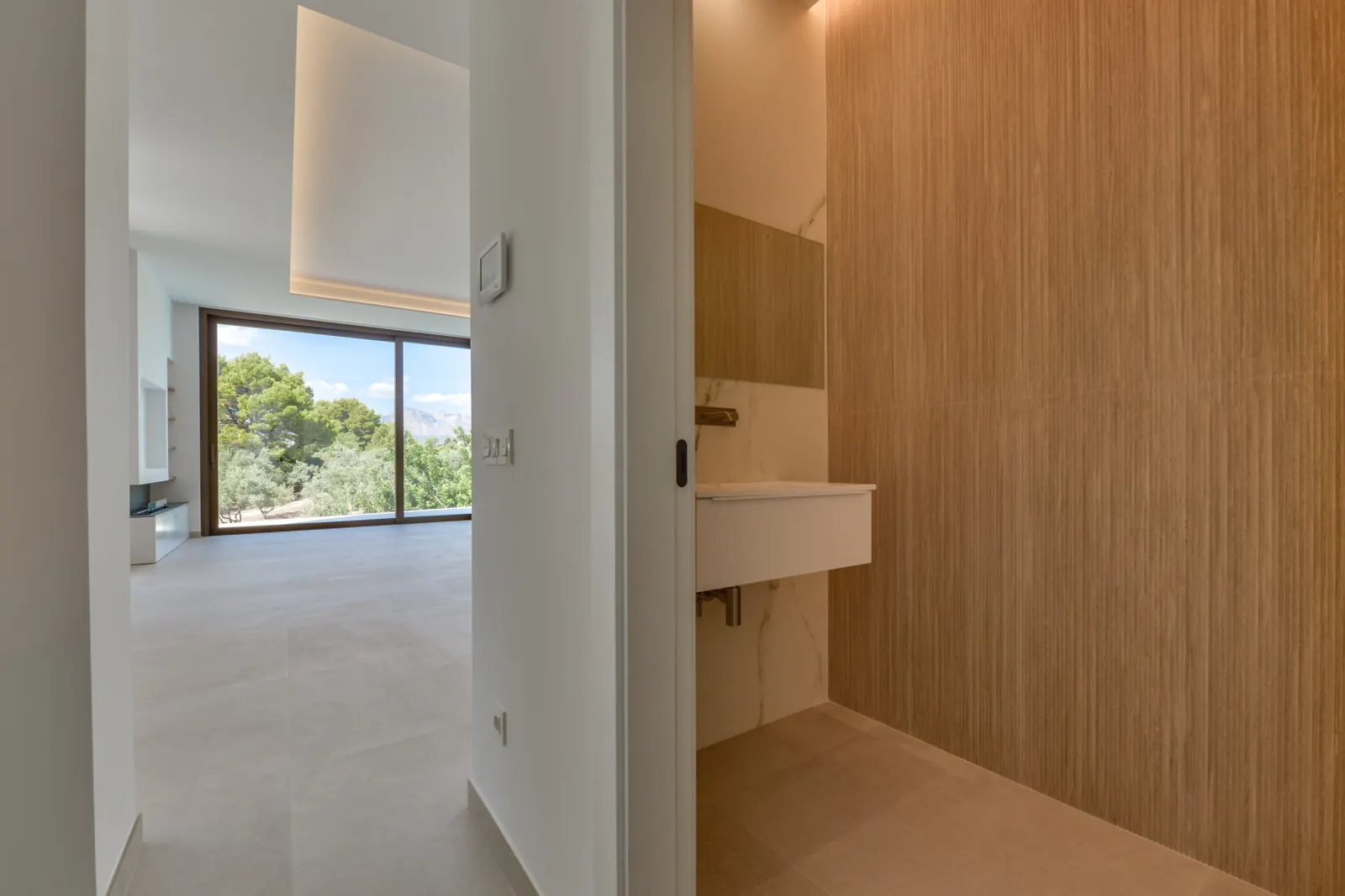 El aseo de cortesía se reviste de madera de veta continua, en contraste con el mármol del lavabo suspendido y con la luz rasante que baja por la pared.