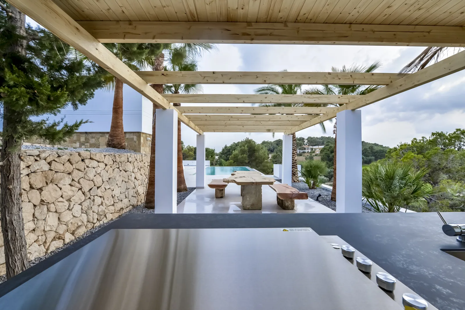 La cocina exterior de acero inoxidable mira al comedor de verano bajo la pérgola, con la mesa de tablón macizo sobre pies de piedra y la piscina al fondo.