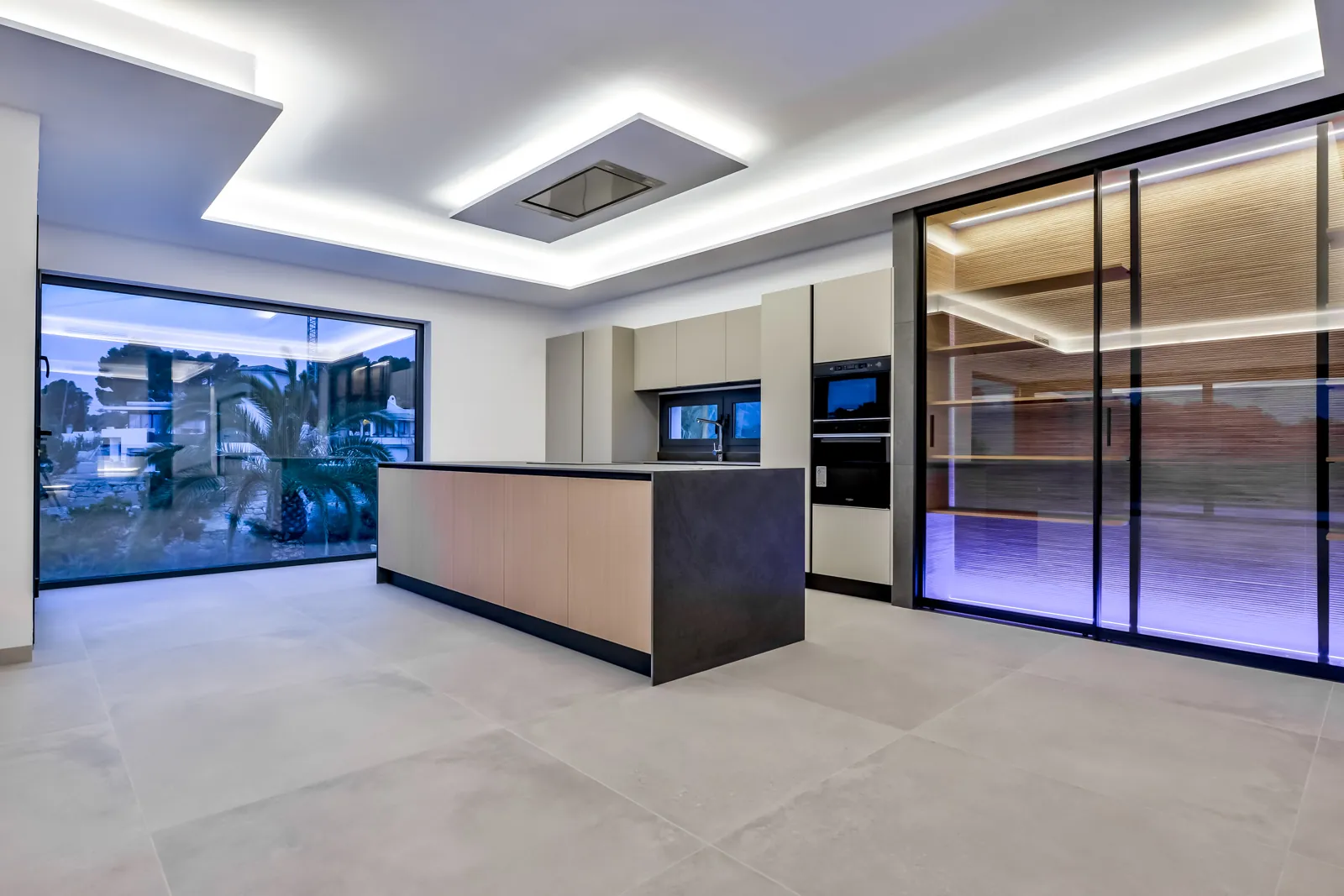 La cocina interior, con la isla de frente de madera y encimera de piedra oscura. La luz perimetral del techo evita las sombras duras sobre el plano de trabajo.