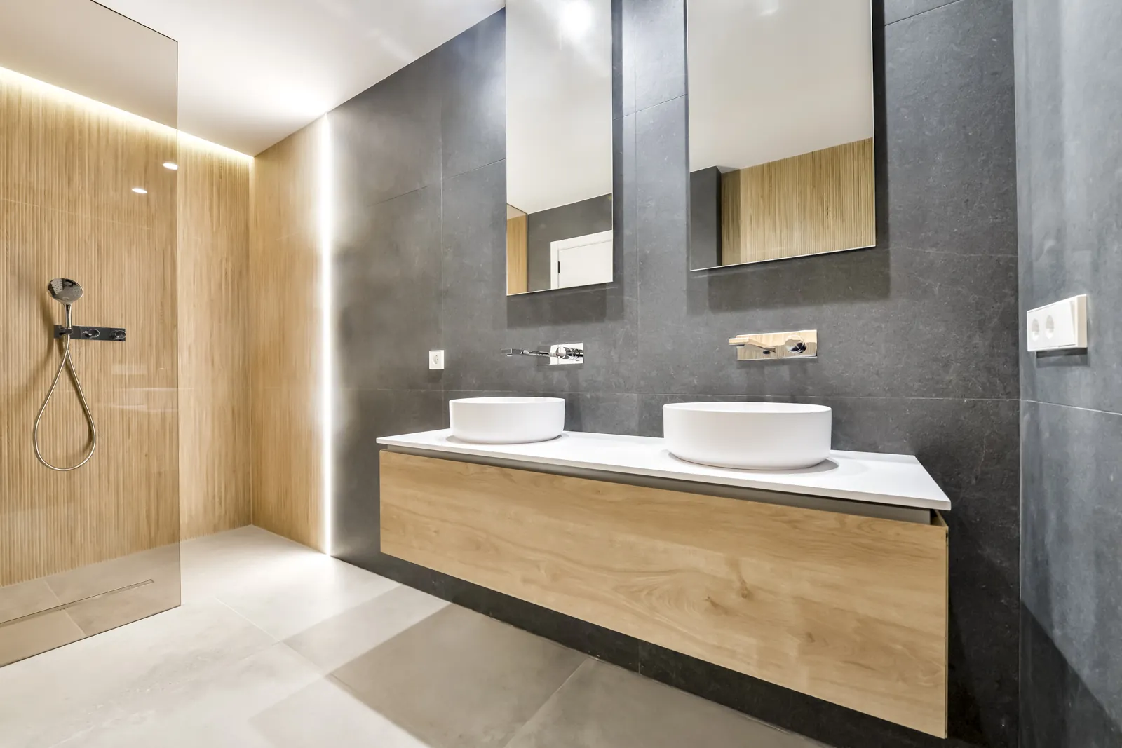 Baño principal: dos lavabos sobre un mueble suspendido de roble, frente de piedra gris y ducha corrida revestida de madera de veta vertical.