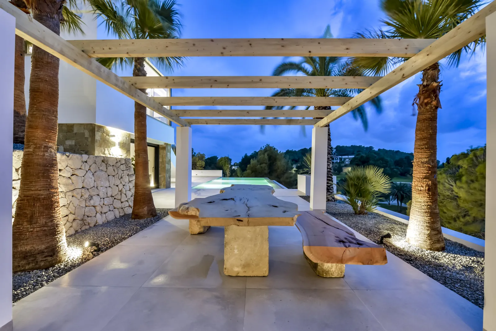 La pérgola al anochecer. La luz enterrada lava los troncos de las palmeras y el muro de piedra seca, y deja la mesa y los bancos de tablón en penumbra.