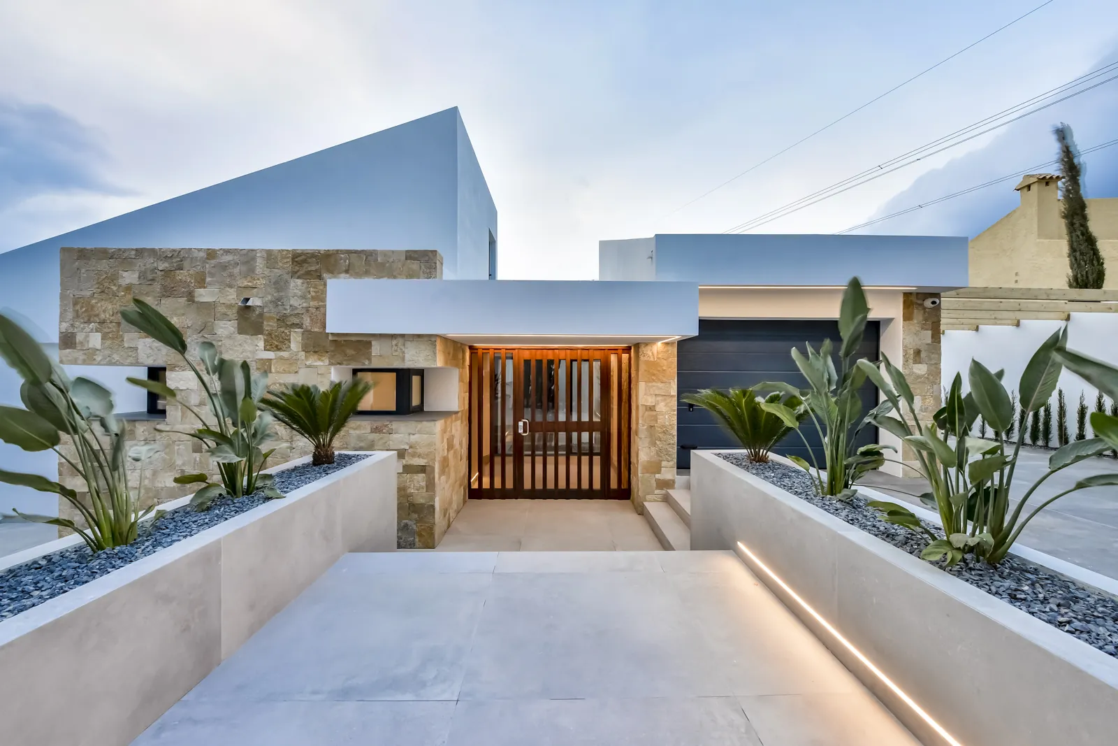 El acceso: la puerta de listones de madera queda retroiluminada entre los paños de piedra, con las jardineras de gravas y estrelitzias acompañando la rampa.