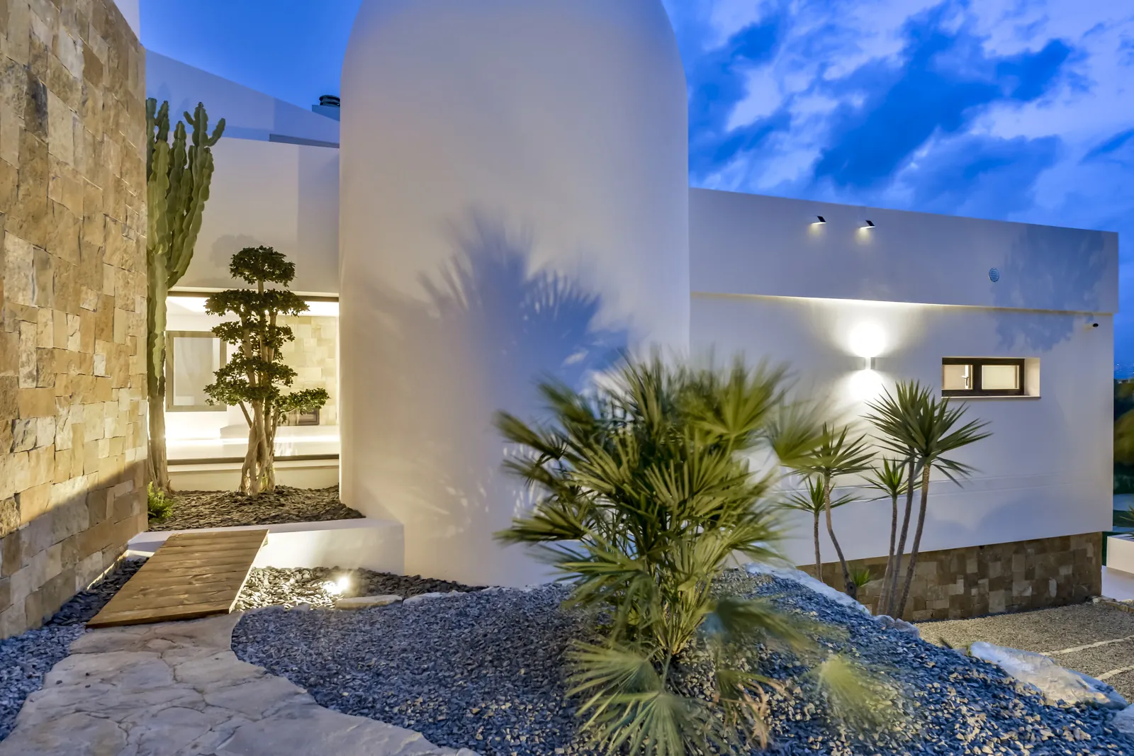 El patio de acceso de noche, con el volumen curvo encalado en el centro. Una pasarela de madera cruza las gravas hasta el porche, entre cactus y un olivo podado.