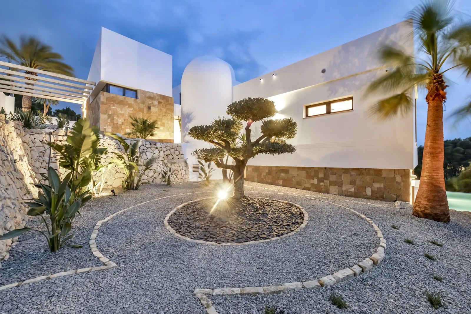 El olivo centenario ocupa el centro del patio, sobre un círculo de grava perfilado con adoquín. Al fondo, el zócalo de piedra dorada de la vivienda.