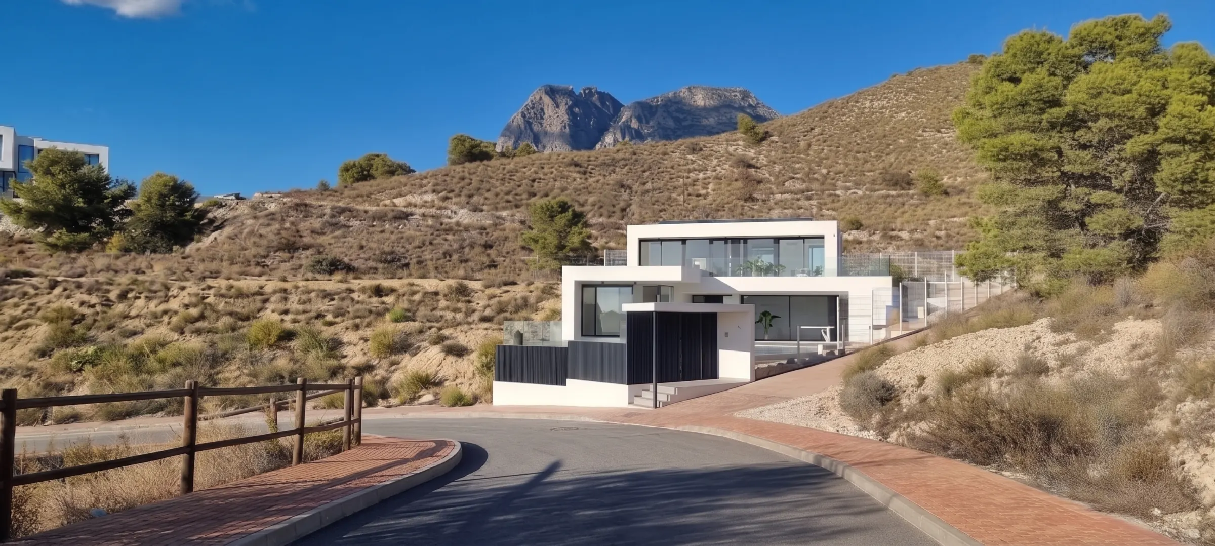La casa desde la curva del vial, encajada en la ladera de matorral. Al fondo, el perfil hendido del Puig Campana sobre Finestrat.