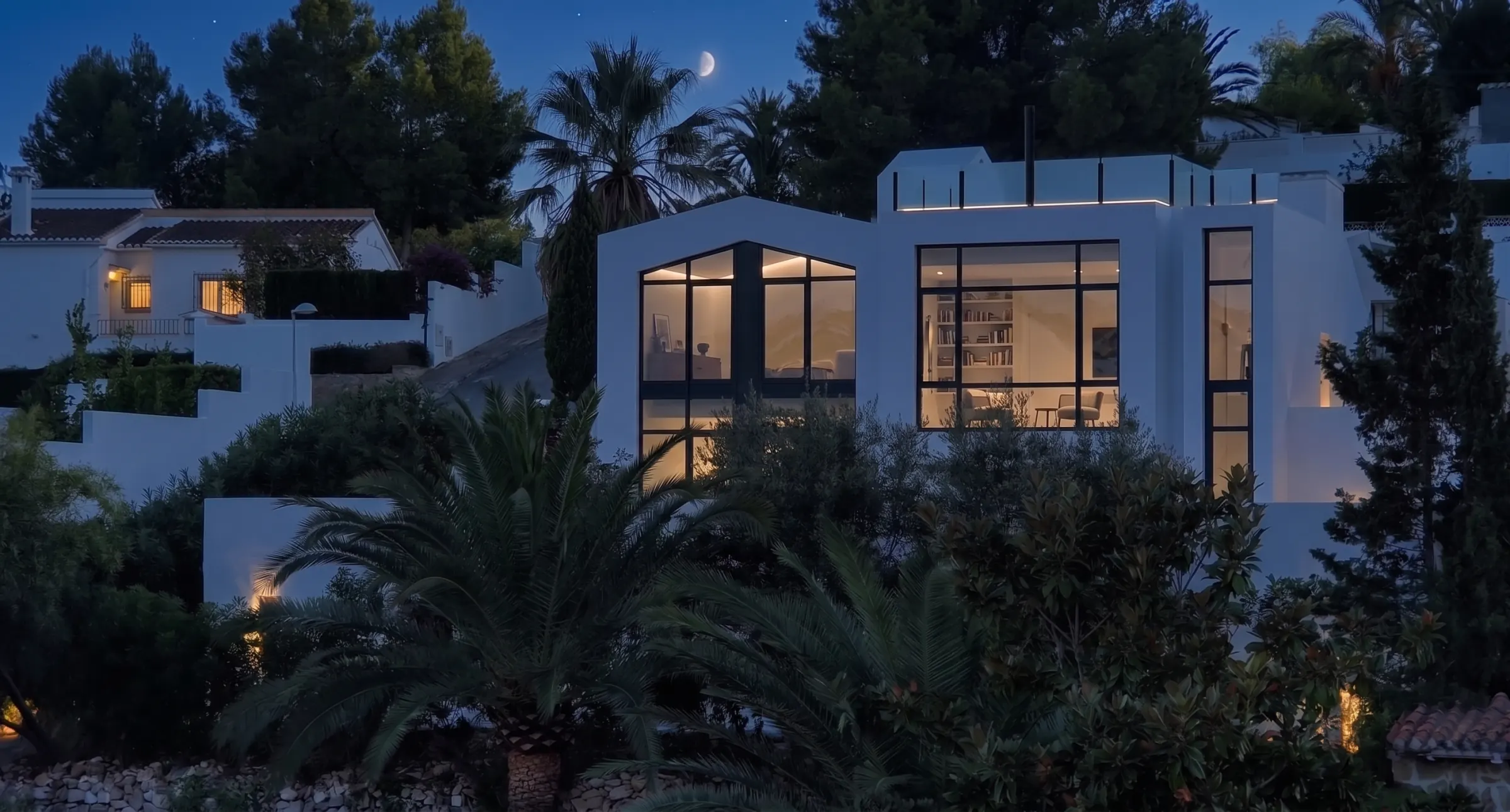 La casa de noche entre la vegetación. El perfil a dos aguas y los grandes ventanales de carpintería negra distinguen la rehabilitación de las villas de teja del entorno.