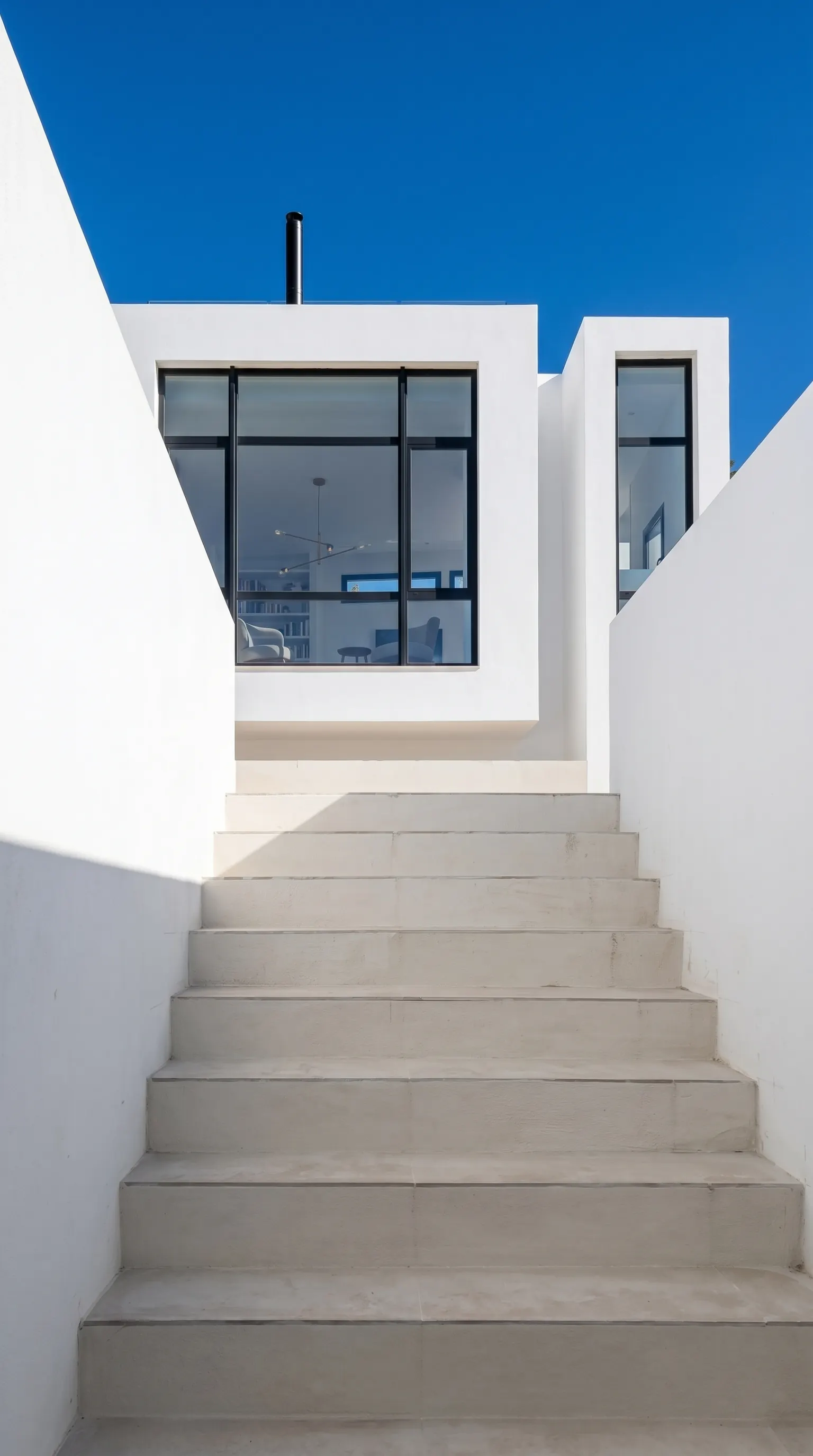 La escalera de acceso, con el ventanal del estar recortado contra el cielo. El blanco de los muros y el negro de la carpintería son toda la paleta del exterior.