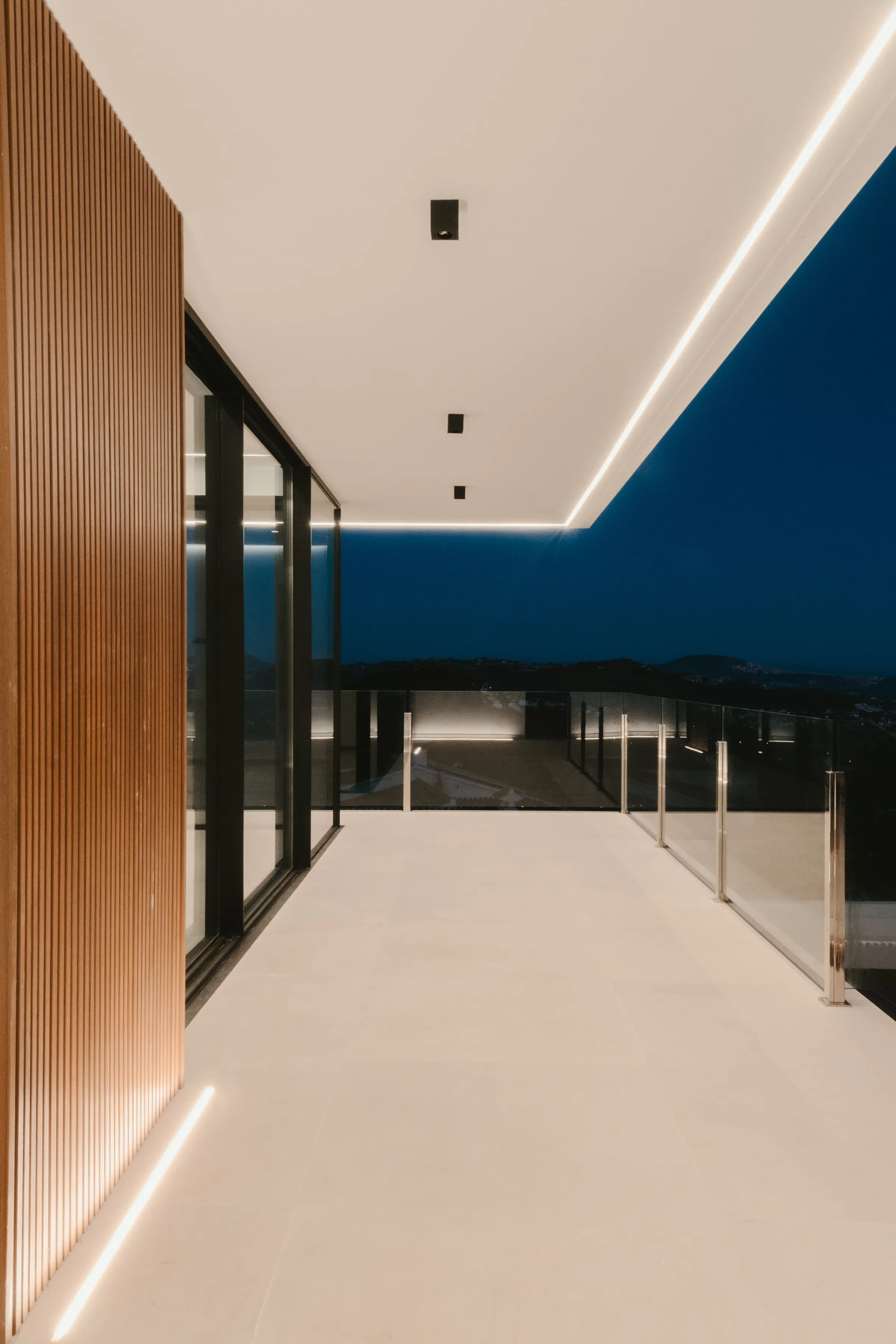 El paso cubierto que recorre la fachada, entre el paño de listones de madera y la barandilla de vidrio. Una línea de luz continua marca el encuentro del techo con el exterior.