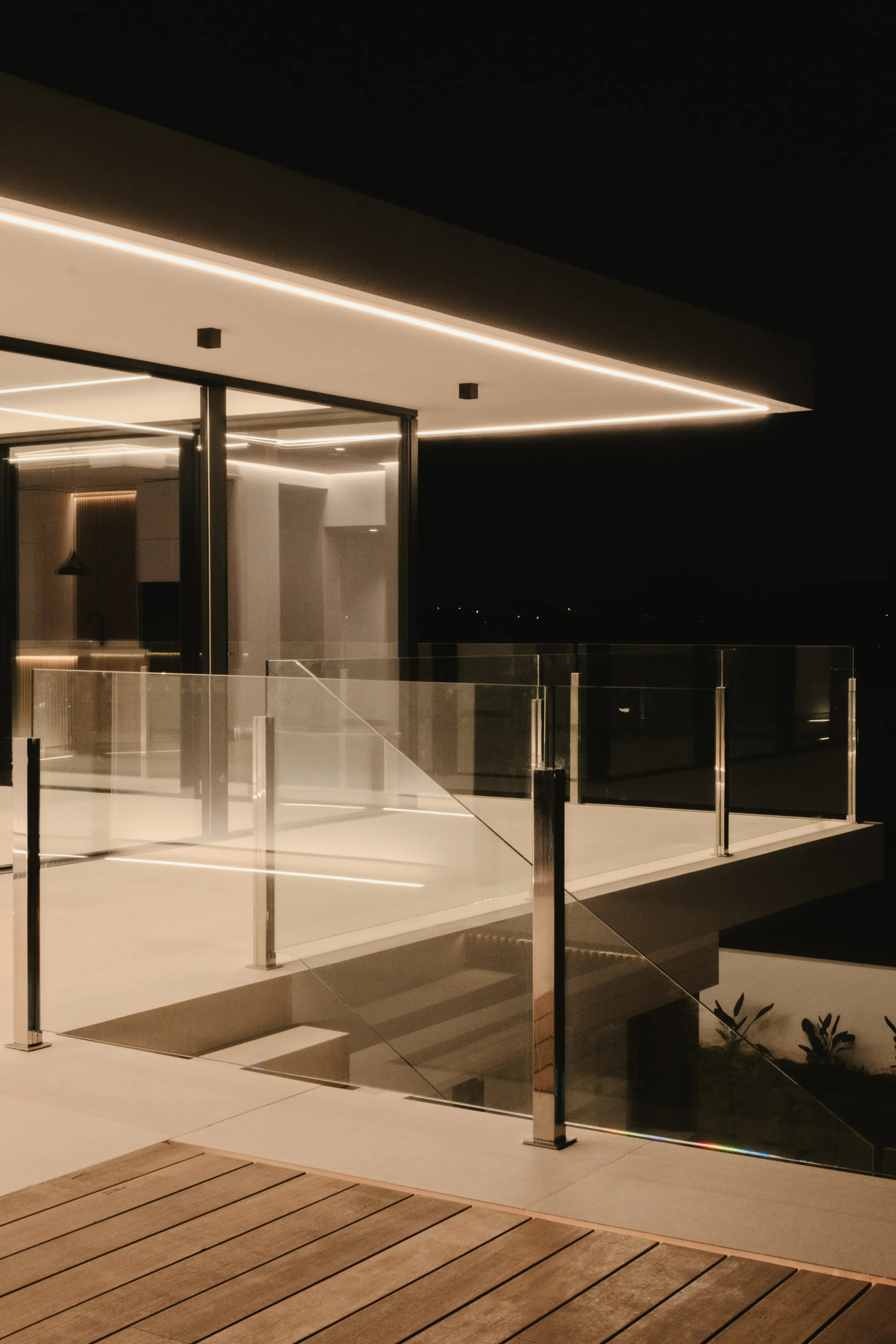 La terraza de noche. El voladizo se despega del suelo con una línea de luz en su canto, y el vidrio de la barandilla deja la vista libre desde dentro.